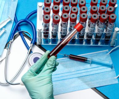 Analityk laboratoryjny trzyma w dłoni fiolkę z krwią pacjenta, w tle położone są różne akcesoria medyczne. /Źródło: 123rf.com