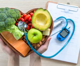 Lekarz trzyma w dłoni tace z warzywami, za którą położone są na stole glukometr, stetoskop i notes. /Źródło: 123rf.com