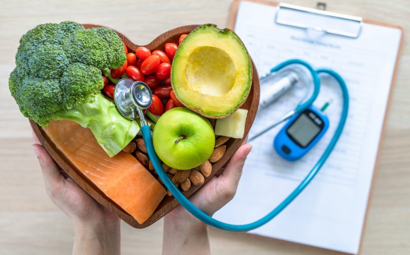 Lekarz trzyma w dłoni tace z warzywami, za którą położone są na stole glukometr, stetoskop i notes. /Źródło: 123rf.com