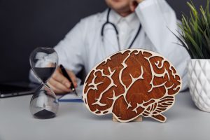 Lekarz neurolog przeprowadza konsultacje, na biurku przed nim leży drewniany model mózgu oraz klepsydra. /Źródło: 123rf.com