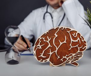 Lekarz neurolog przeprowadza konsultacje, na biurku przed nim leży drewniany model mózgu oraz klepsydra. /Źródło: 123rf.com