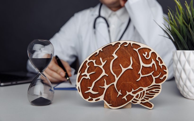 Lekarz neurolog przeprowadza konsultacje, na biurku przed nim leży drewniany model mózgu oraz klepsydra. /Źródło: 123rf.com