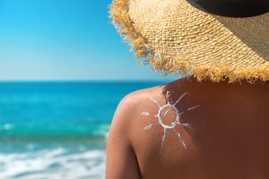 Kobieta ma posmarowane plecy kremem z filtrem, ma założony kapelusz i przebywa na pięknej plaży. /Źródło: 123rf.com