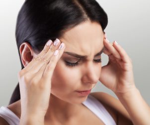Młoda kobieta w długich, ciemnych włosach trzyma się dłońmi za skronie, ponieważ boli ją mocno głowa. Źródło: 123rf.com