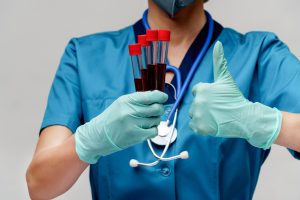 Pielęgniarz odziany w niebieski, sterylny ubiór medyczny trzyma w jednej dłoni probówki z pobraną wcześniej pacjentom krwią, a drugą dłonią pokazuje gest kciuka w górę. Źródło: 123rf.com