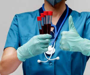 Pielęgniarz odziany w niebieski, sterylny ubiór medyczny trzyma w jednej dłoni probówki z pobraną wcześniej pacjentom krwią, a drugą dłonią pokazuje gest kciuka w górę. Źródło: 123rf.com