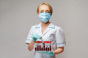 Pielęgniarka w pozycji stojącej ubrana w strój medyczny z maseczką ochronną na twarzy trzyma w dłoniach  fiolki z pobranymi próbkami krwi do celu diagnostyki laboratoryjnej. Źródło: 123rf.com