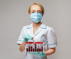Pielęgniarka w pozycji stojącej ubrana w strój medyczny z maseczką ochronną na twarzy trzyma w dłoniach fiolki z pobranymi próbkami krwi do celu diagnostyki laboratoryjnej. Źródło: 123rf.com