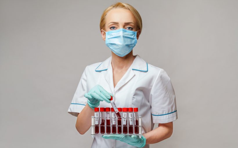 Pielęgniarka w pozycji stojącej ubrana w strój medyczny z maseczką ochronną na twarzy trzyma w dłoniach fiolki z pobranymi próbkami krwi do celu diagnostyki laboratoryjnej. Źródło: 123rf.com