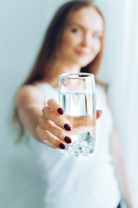 Młoda kobieta wyciąga przed siebie rękę, w której trzyma szklankę wypełnioną wodą. /Źródło: 123rf.com