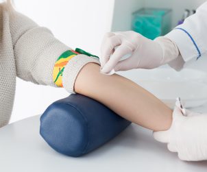 Preparation for blood test with beautiful young blond woman by female doctor in white coat medical uniform on the table in white bright room. Nurse rubbing a hand styryl patient tissue.